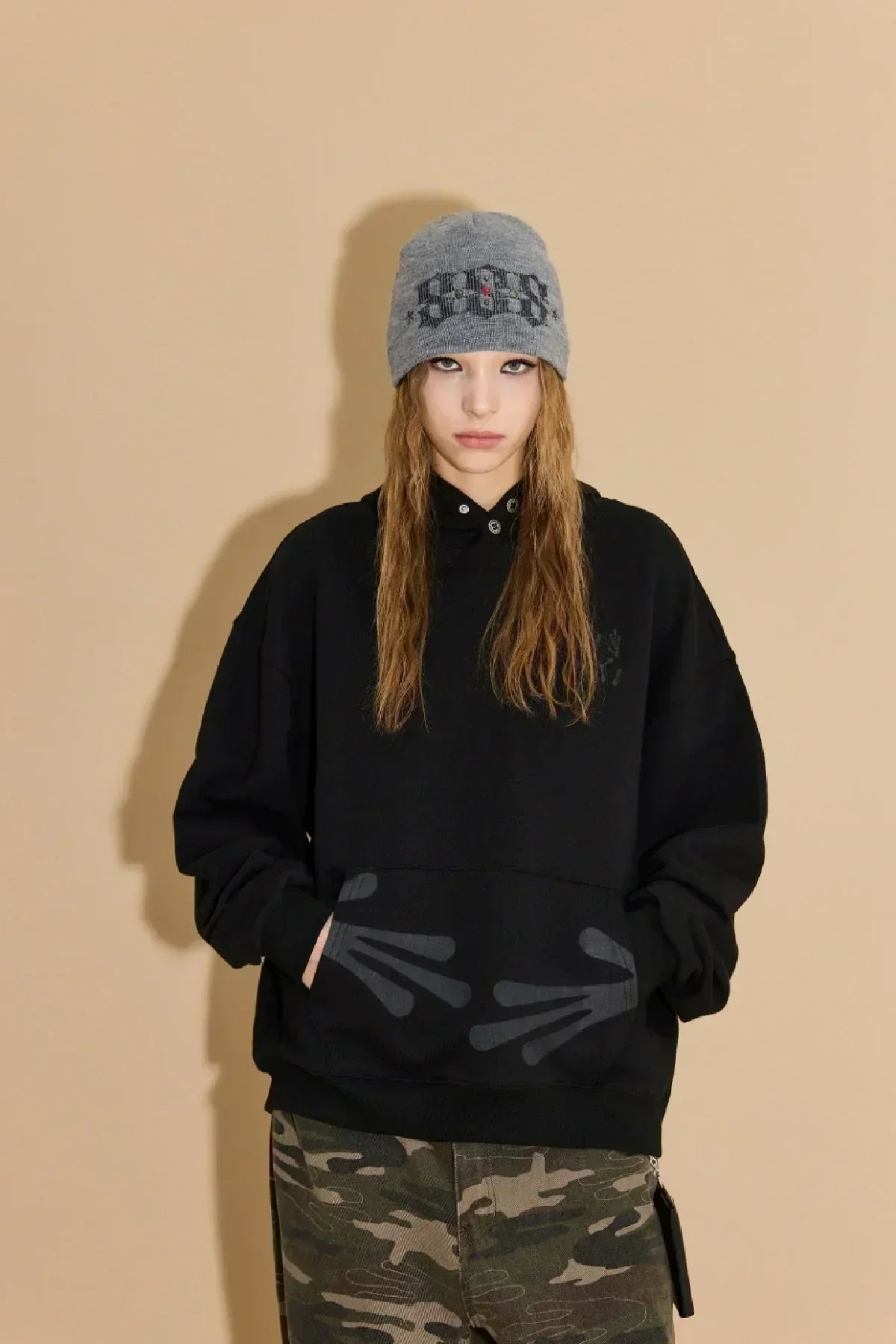 Model wearing black hoodie with alien hand prints and camouflage pants against beige background