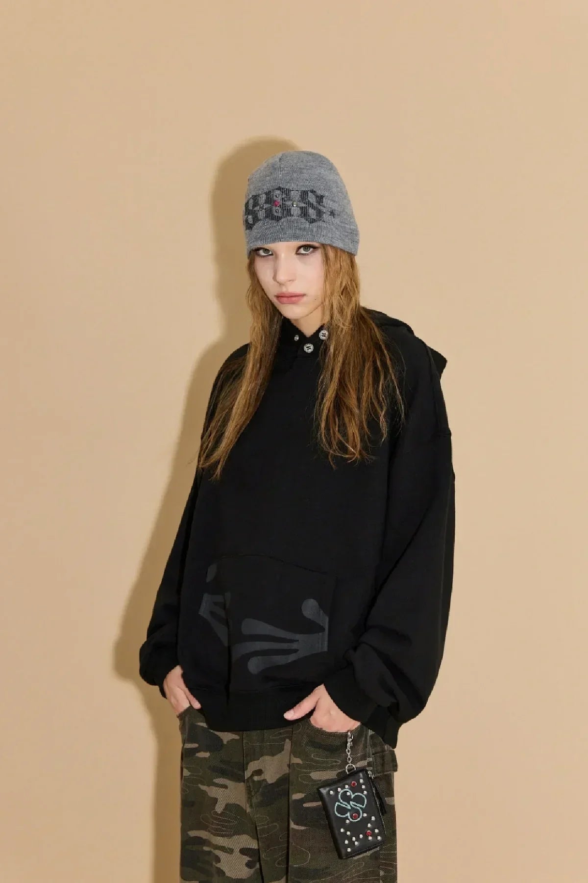 Model wearing black oversized hoodie and camo pants with gray beanie against beige background