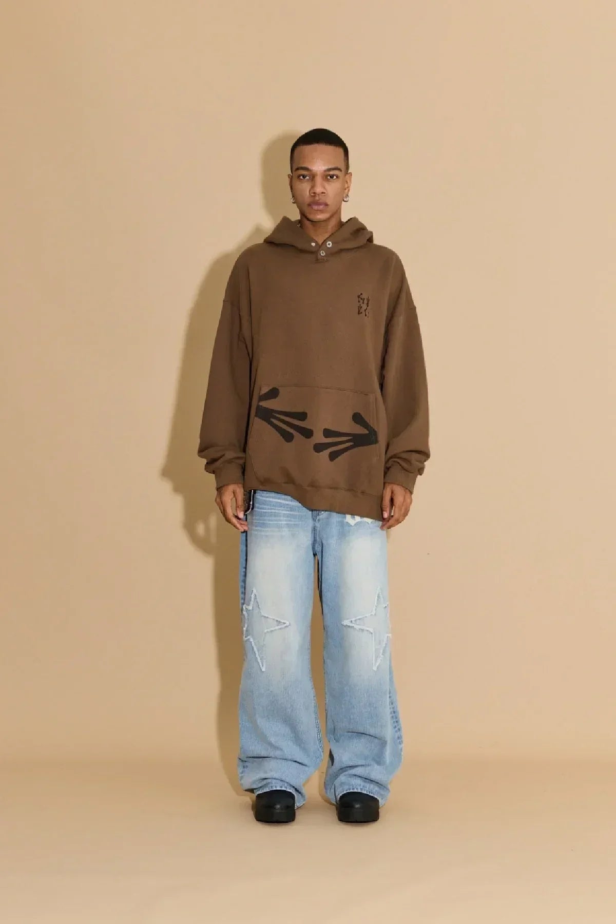 Man wearing brown alien hands hoodie with black graphic arrows and light blue star-patch baggy jeans