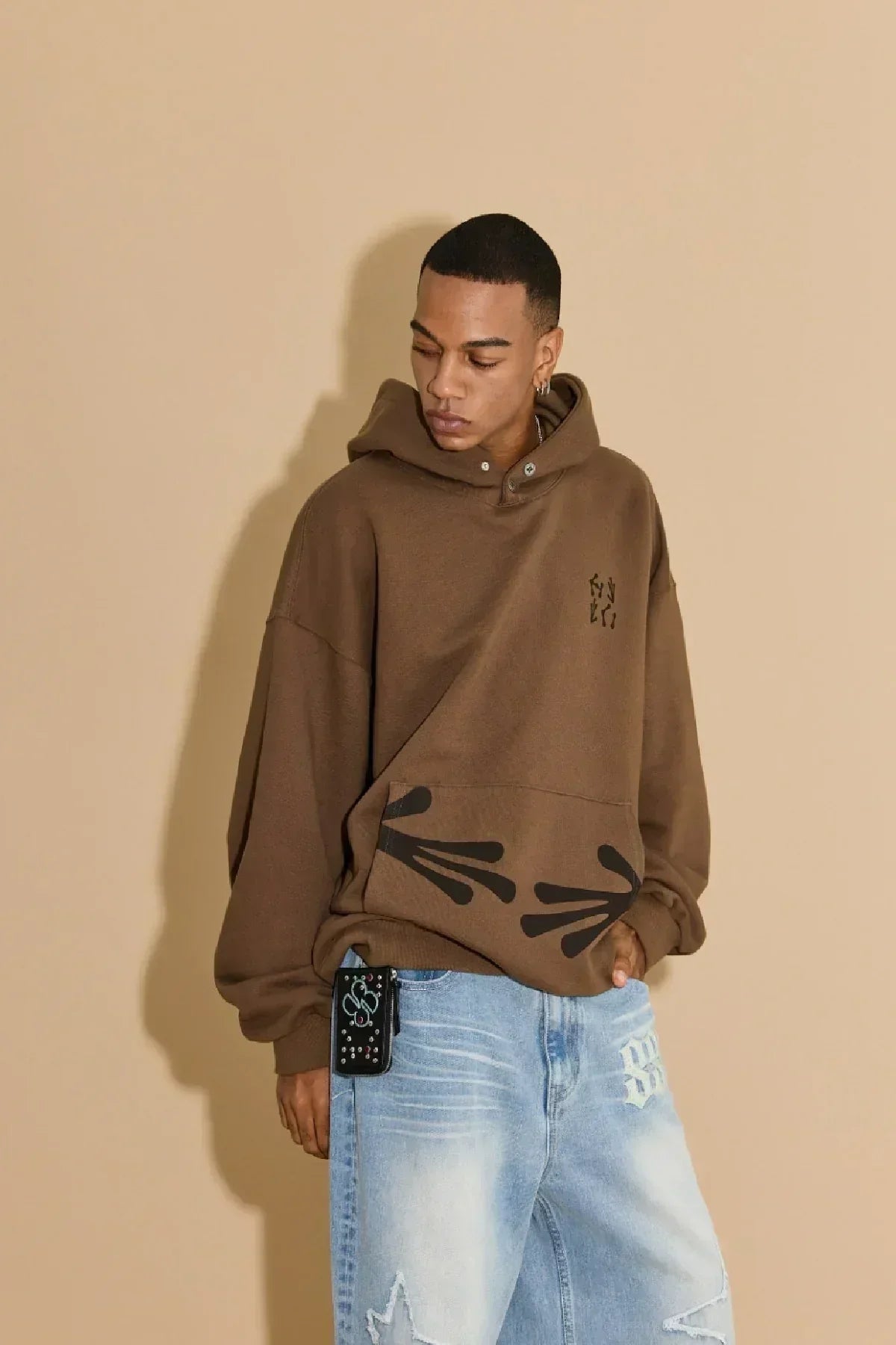 Young man wearing brown hoodie with black alien hand graphics and faded blue jeans against beige background