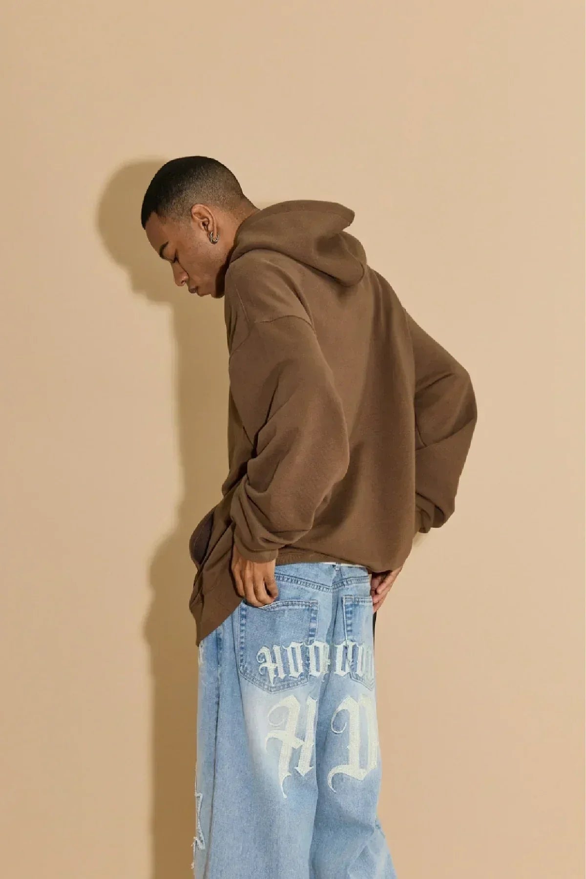 Male model wearing brown hoodie and light blue oversized jeans with white gothic lettering on beige background
