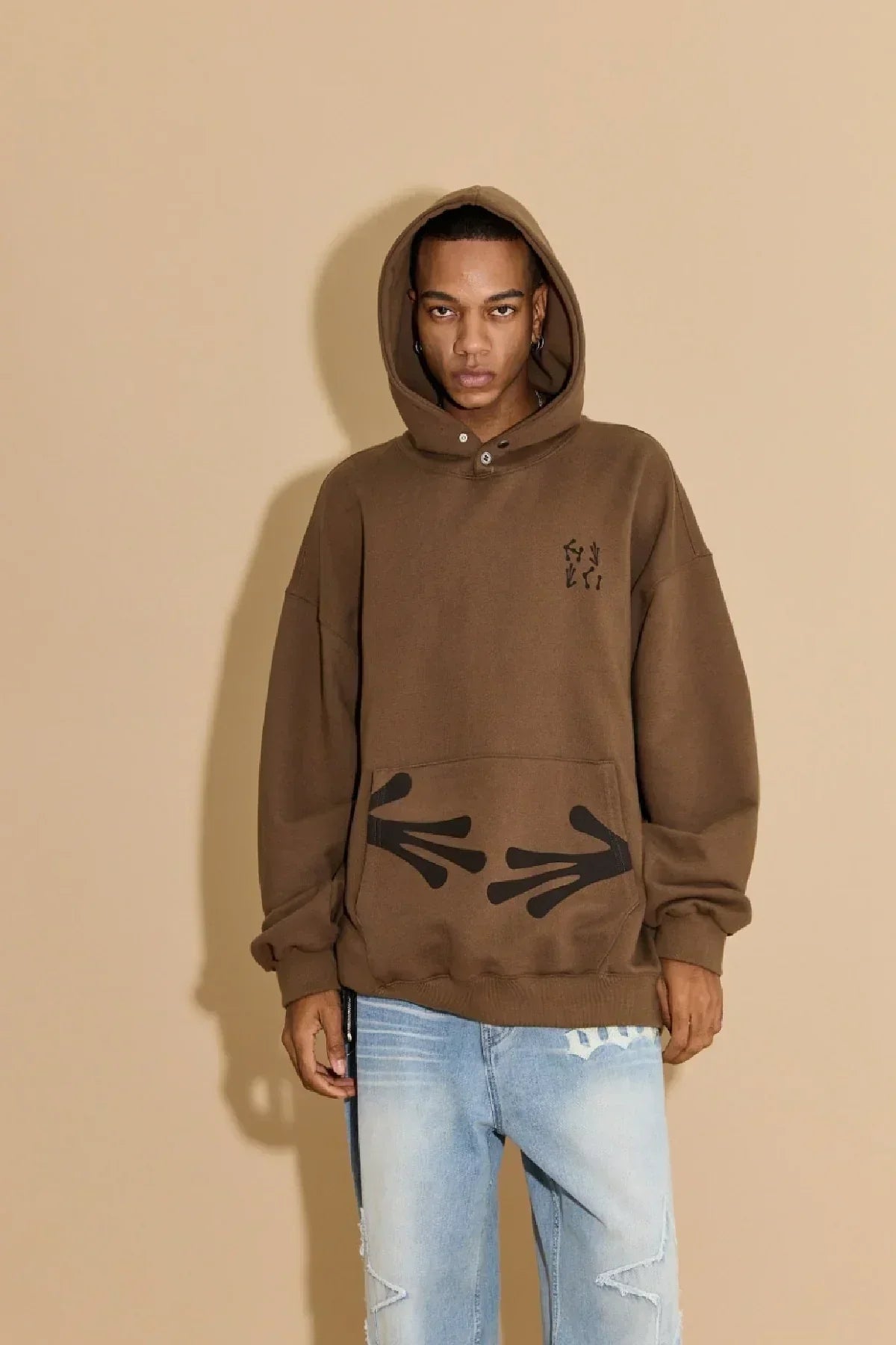 Model wearing brown hoodie with black arrow handprints and light blue ripped jeans against beige background