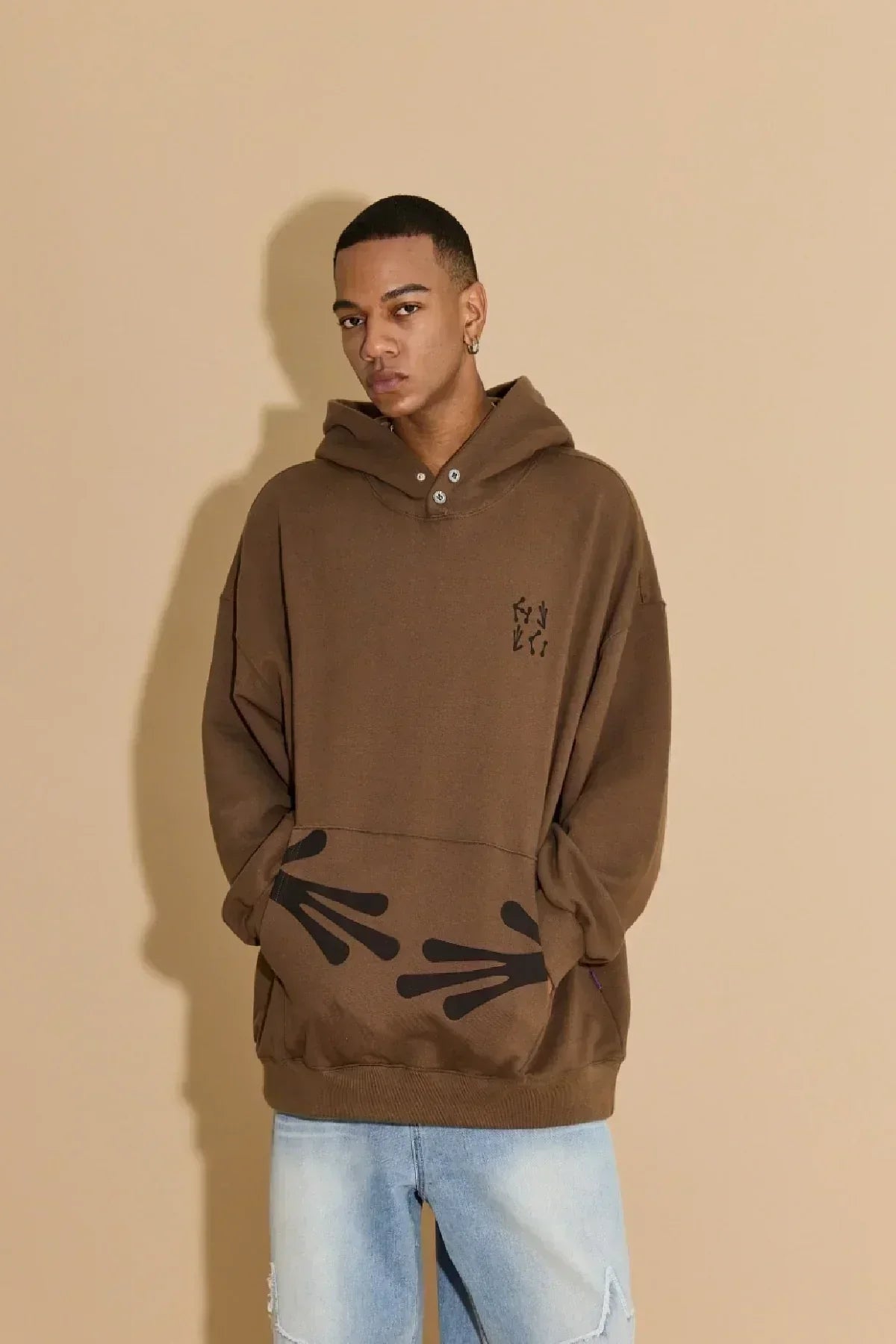 Young man wearing brown hoodie with black alien hands graphic and light blue jeans against beige background