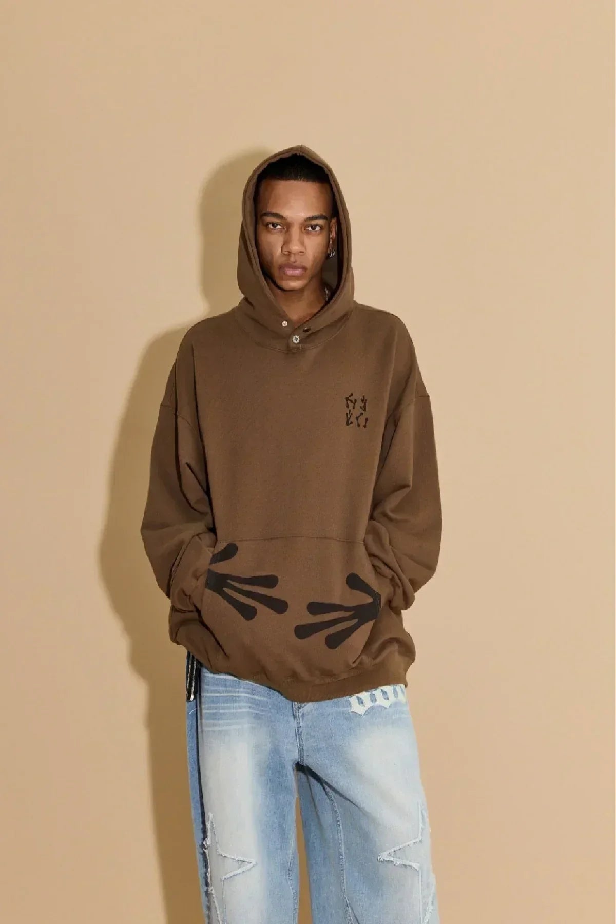 Man wearing brown hoodie with black alien hand prints on front pocket and light blue jeans