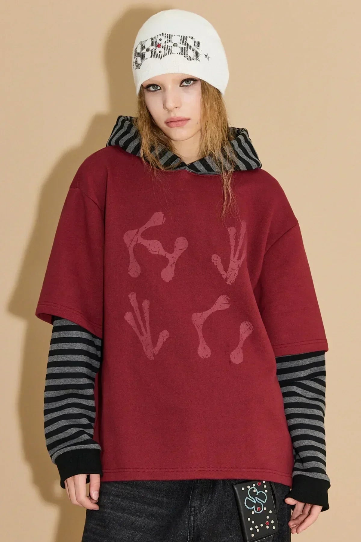 Model wearing burgundy alien-themed layered hoodie with striped black-gray sleeves and white graphic beanie