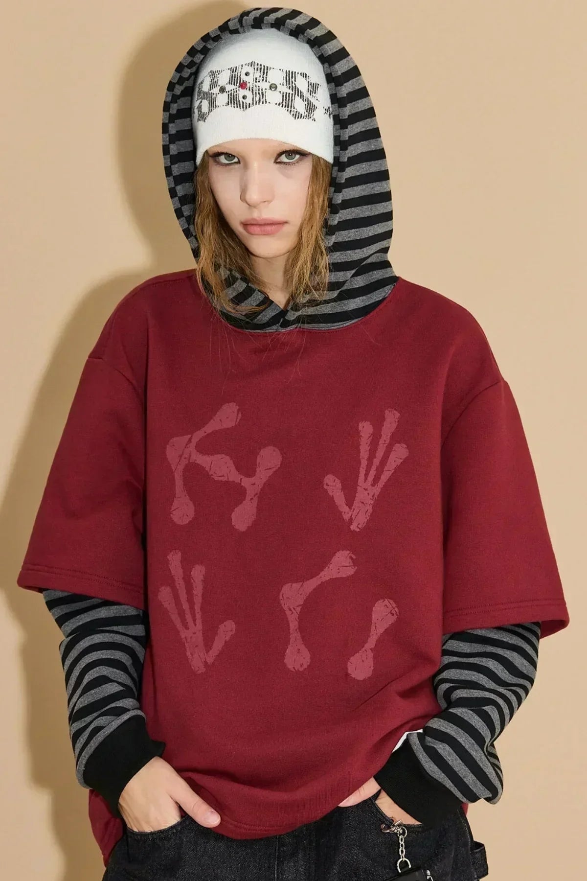 Model wearing burgundy alien hand print hoodie layered over black and gray striped long sleeve shirt with white beanie