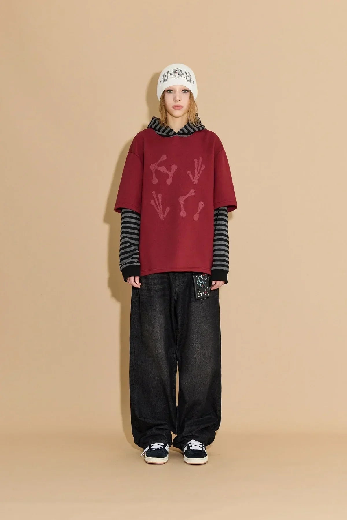 Model wearing burgundy alien hands graphic hoodie layered over striped long sleeves, baggy black jeans, beanie, and sneakers