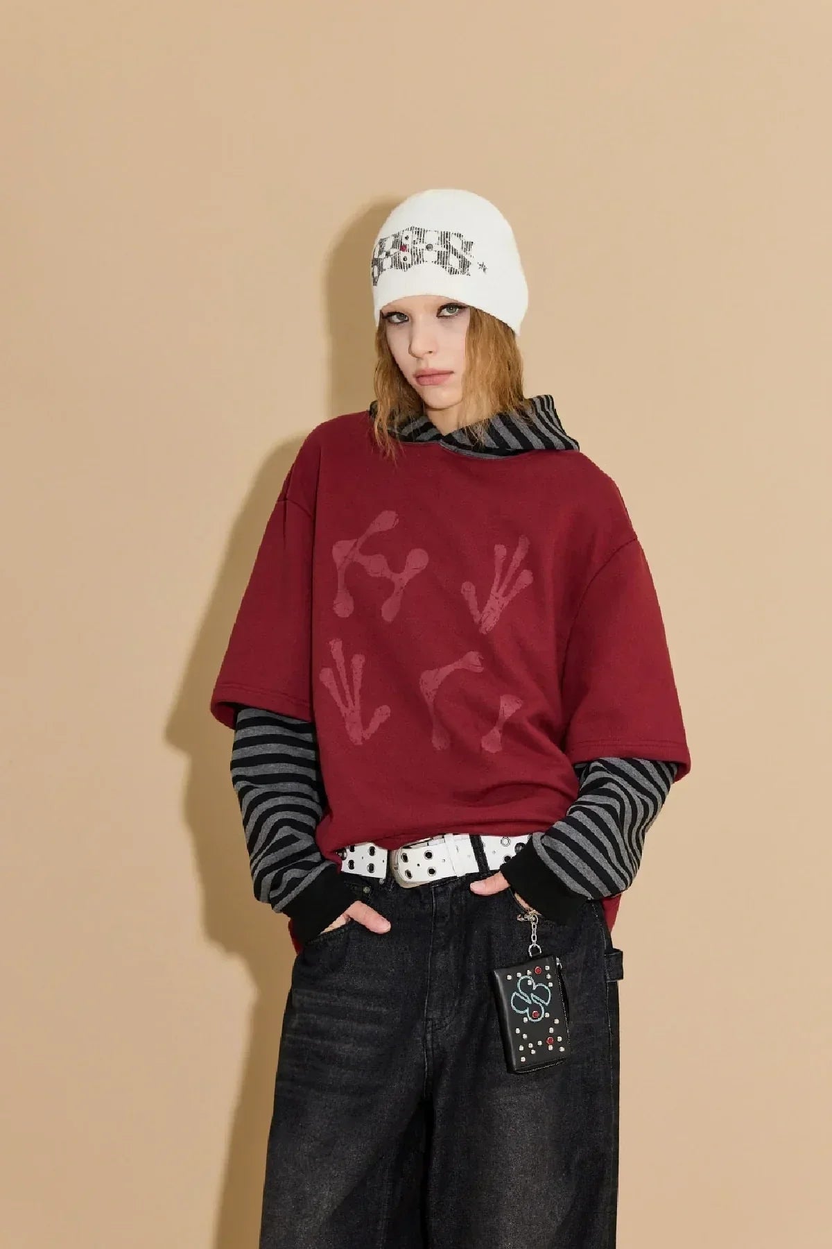 Model in burgundy layered stripe hoodie, black jeans, white belt, and white beanie with graphic text