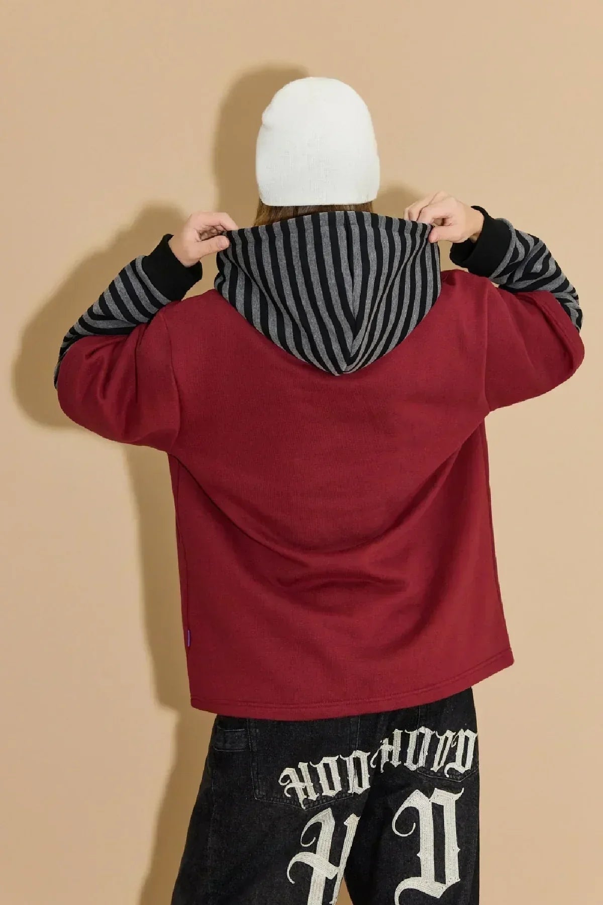 Model wearing burgundy hoodie with black and grey striped hood and sleeves, white beanie, black pants with white lettering