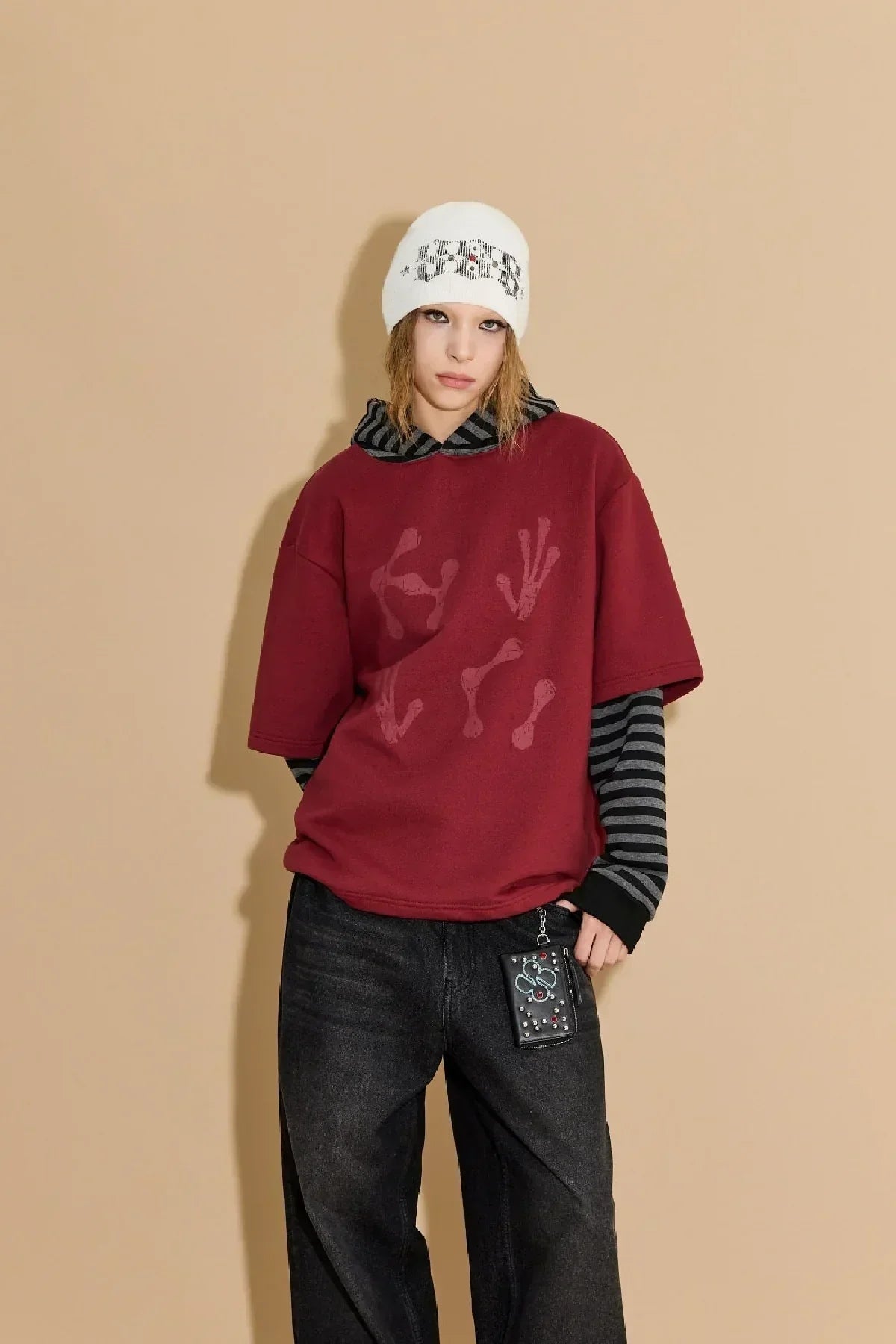 Young person wearing burgundy short-sleeve hoodie over striped long sleeves and a white beanie against beige background