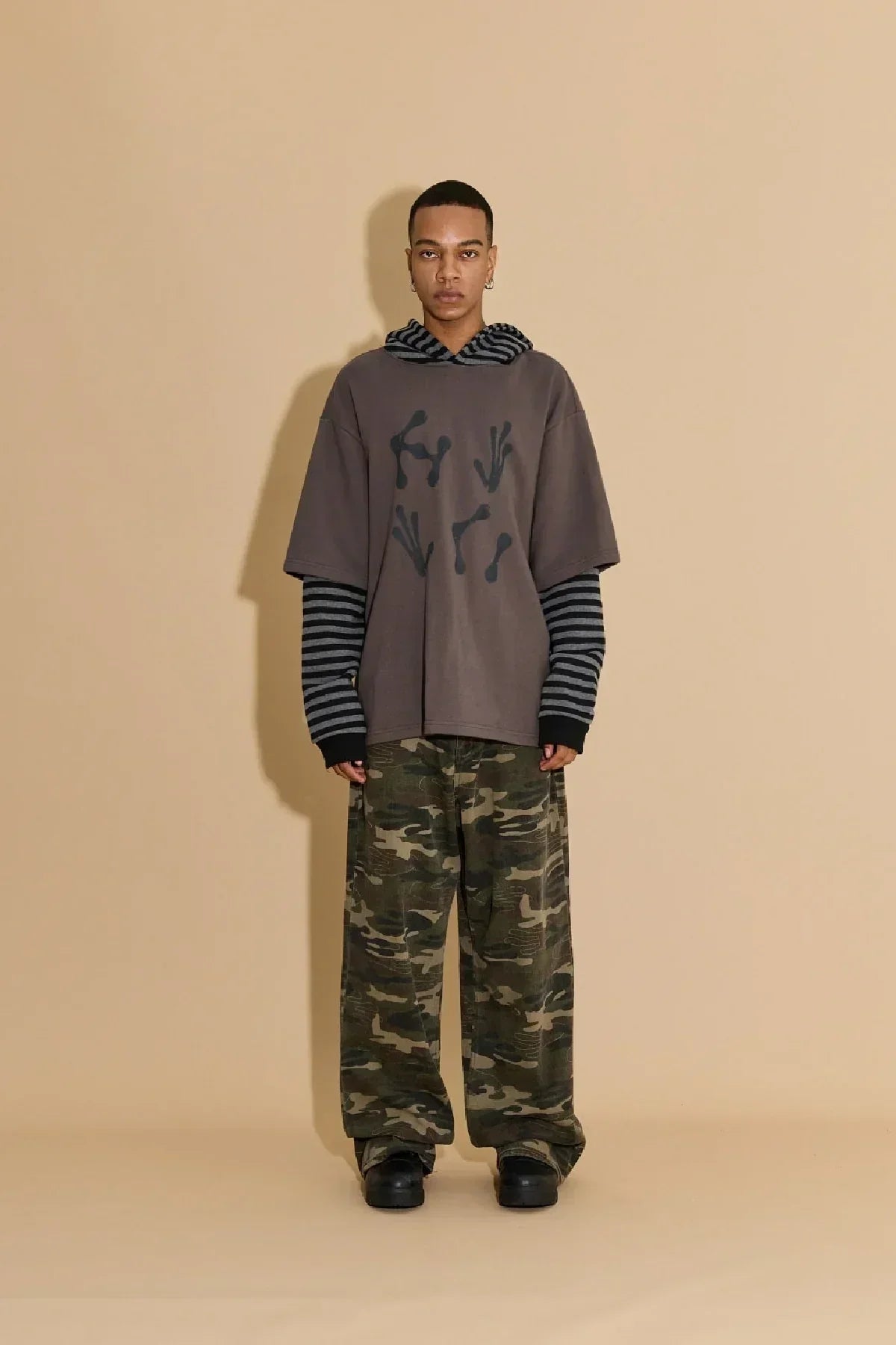 Model wearing charcoal alien hands graphic layered stripe hoodie with camouflage pants and black shoes