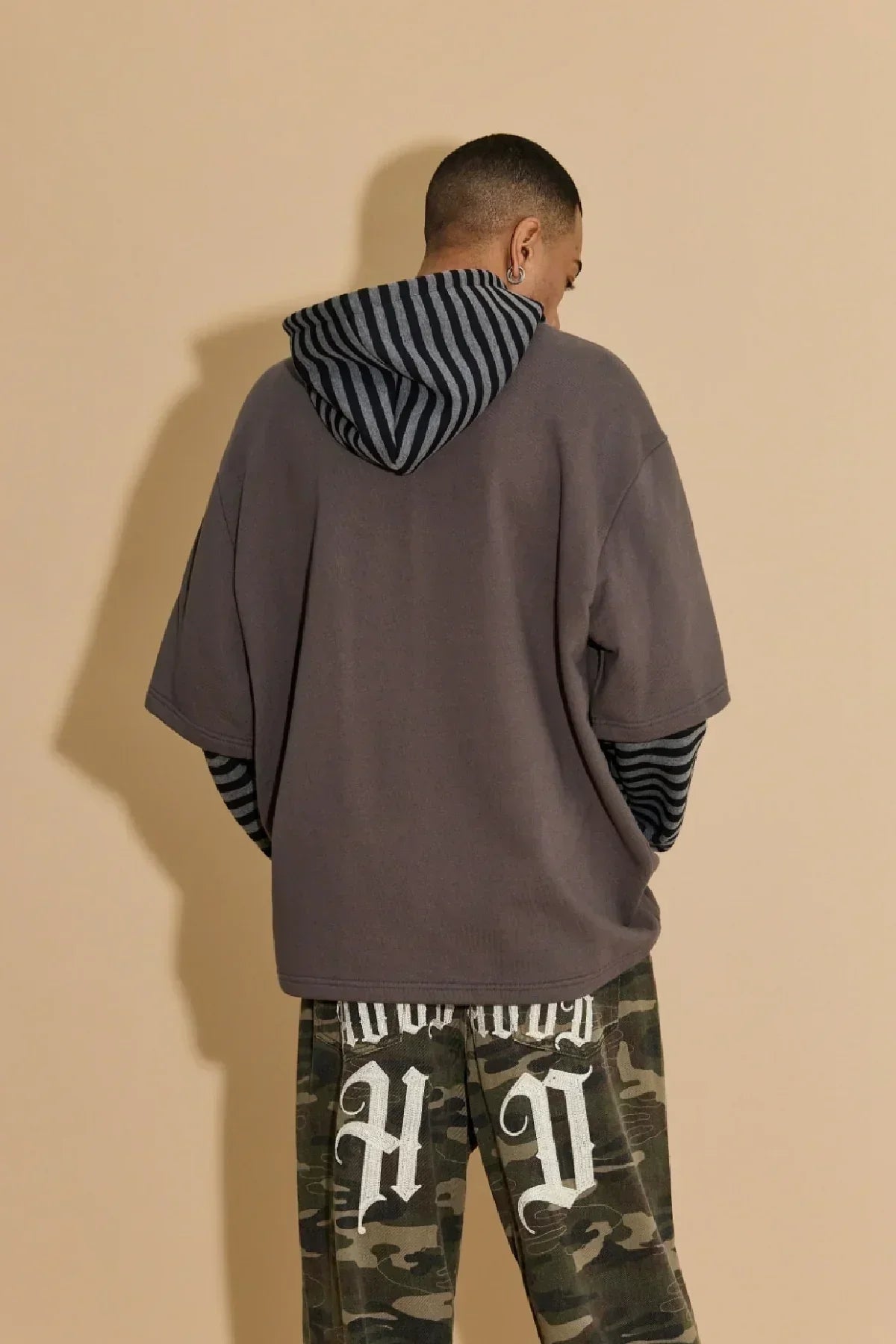 Back view of man wearing charcoal short-sleeve hoodie layered over black and gray striped hood, camouflage pants with white Gothic text