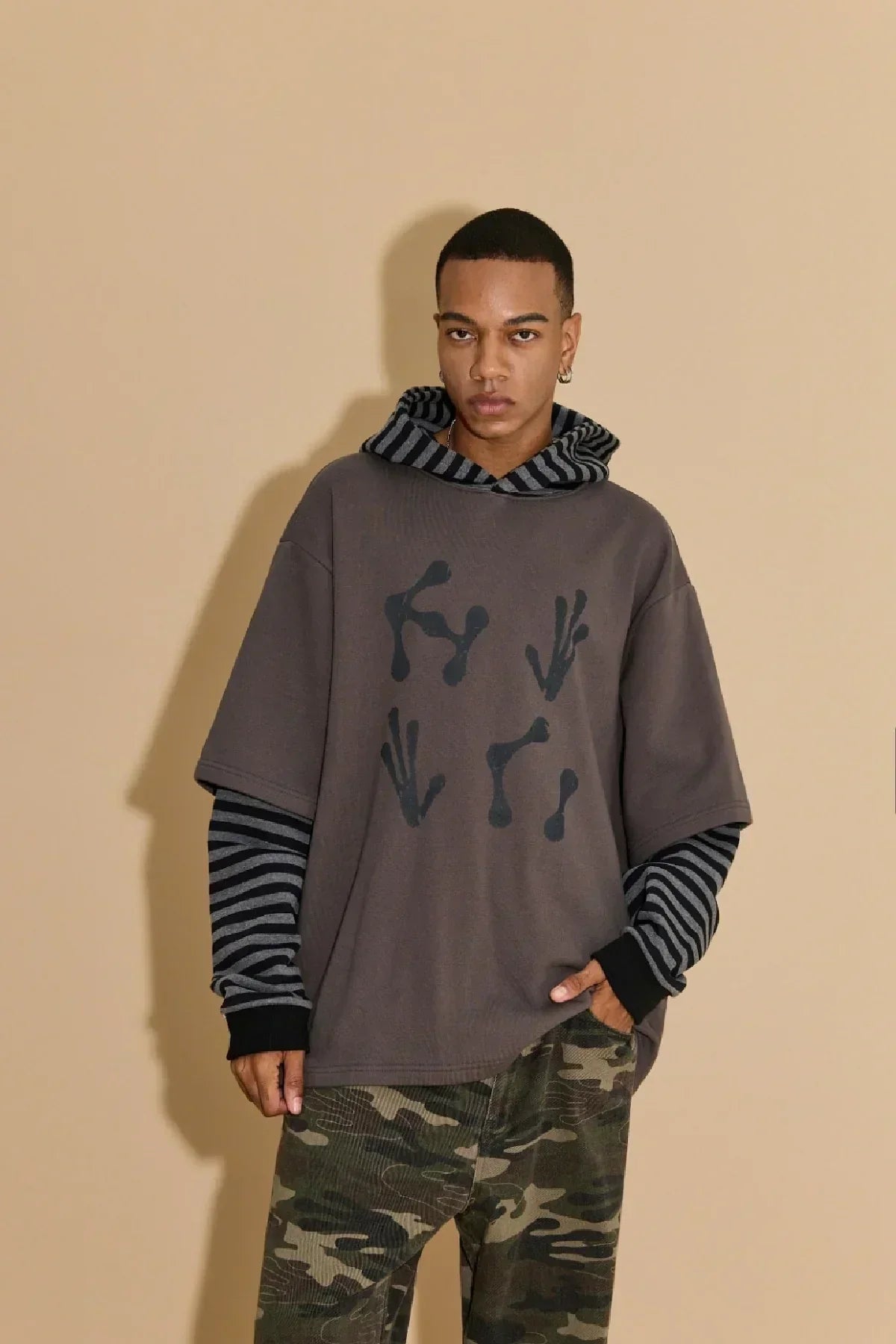 Young man wearing charcoal alien hands print hoodie layered over striped long sleeves and camouflage pants