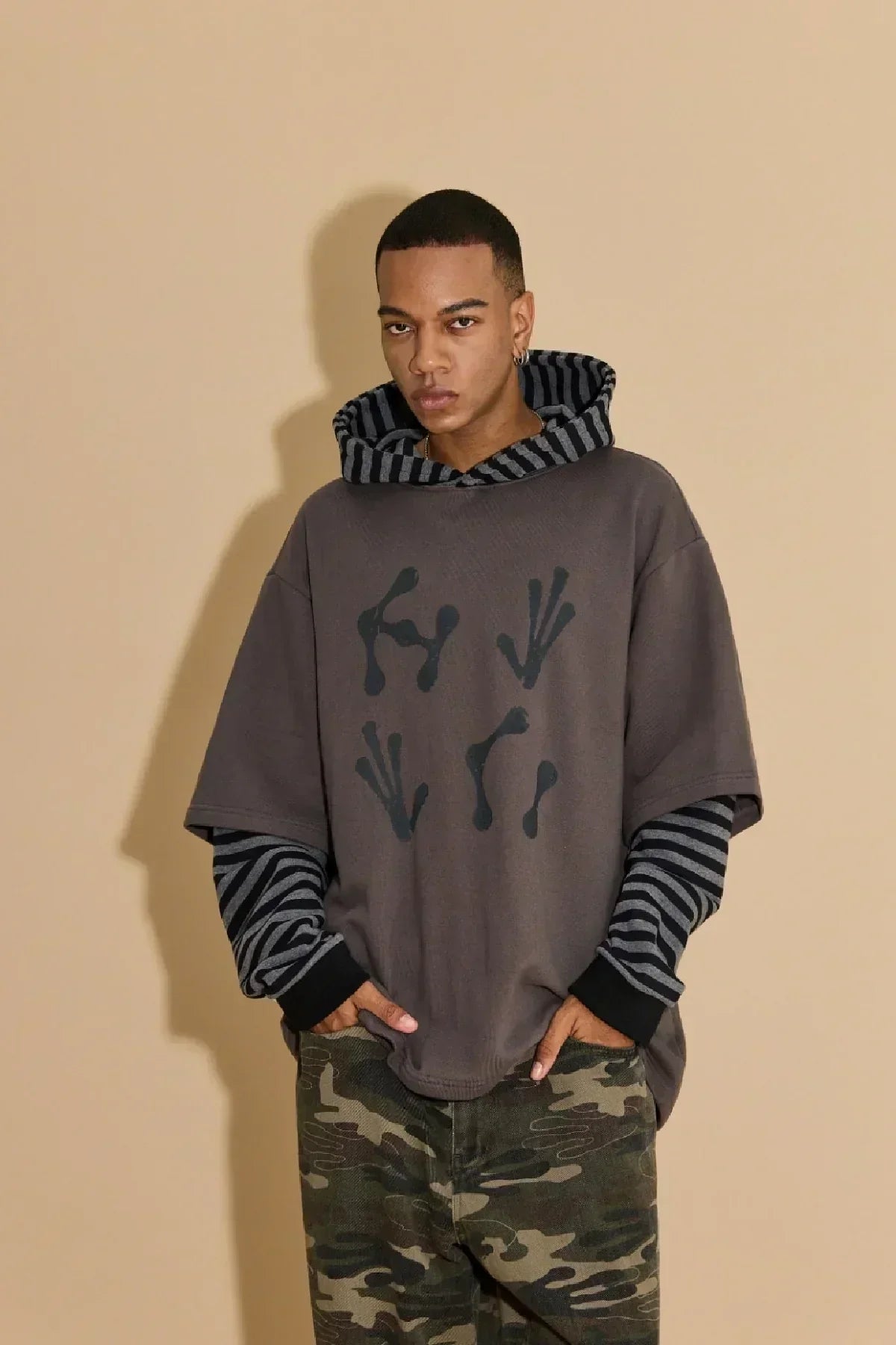 Young man wearing charcoal alien hands layered stripe hoodie and camo pants against beige background