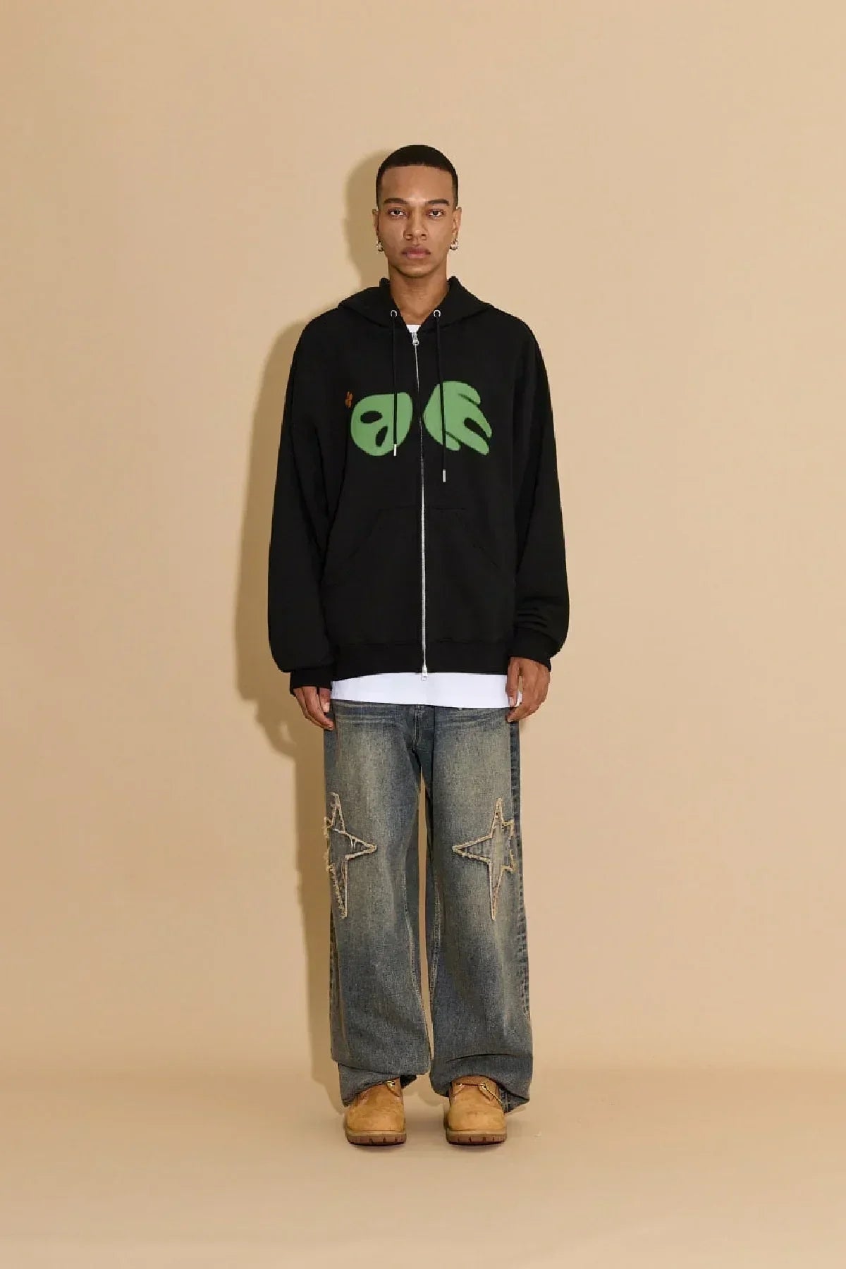 Male model wearing black zip-up hoodie with green alien design, baggy jeans with star patches, tan boots