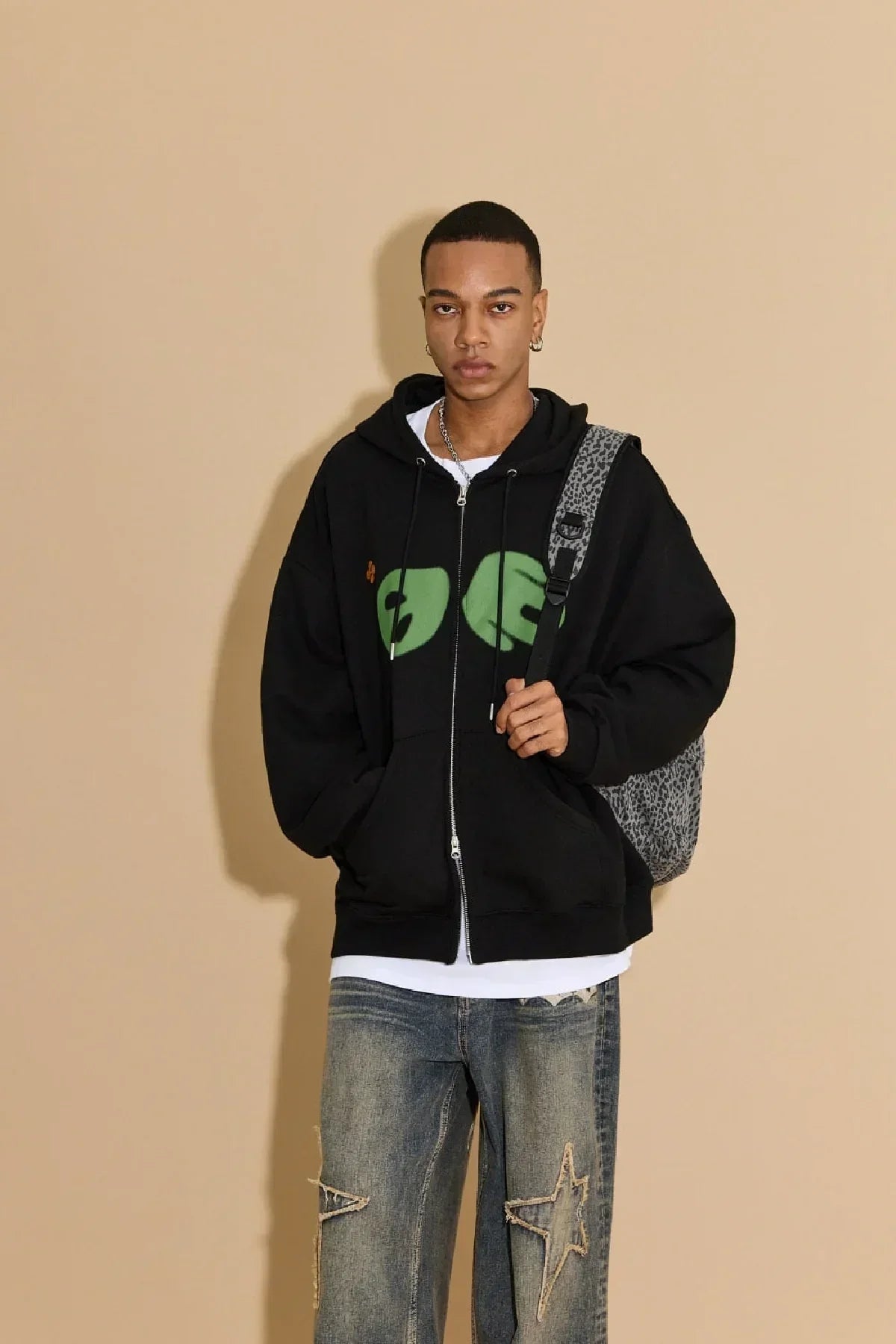 Young man wearing black zip-up hoodie with green alien eyes graphic and star-patched jeans against beige background