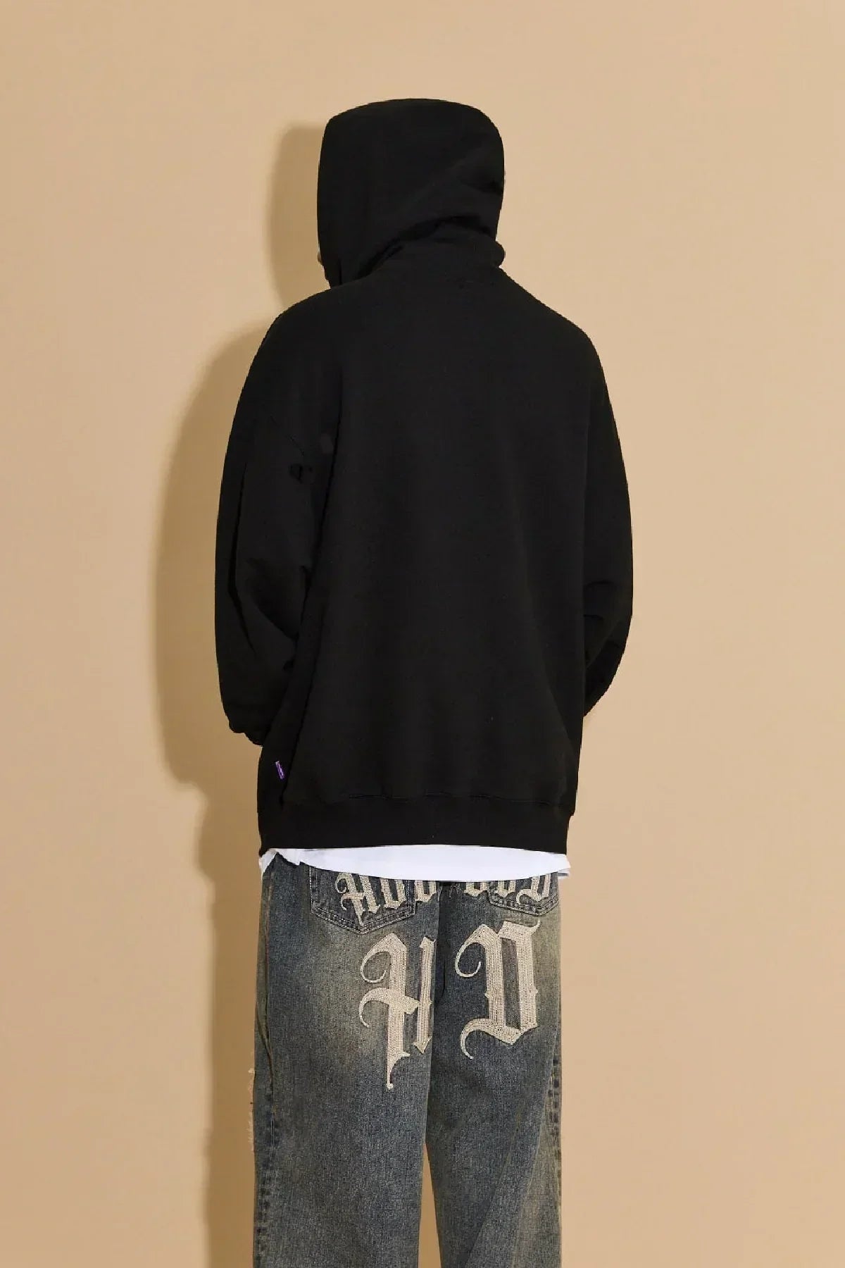Person wearing black hooded zip-up sweatshirt and faded blue jeans with large decorative lettering on back pockets