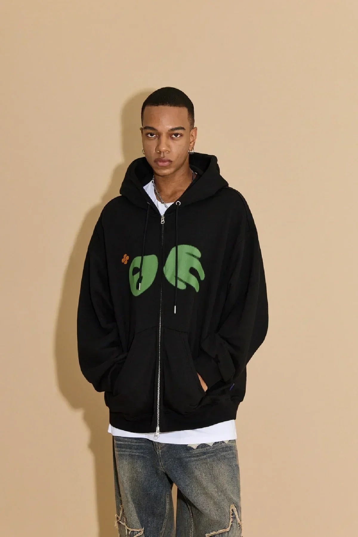 Young man wearing black zip-up hoodie with green abstract design and distressed jeans against beige background