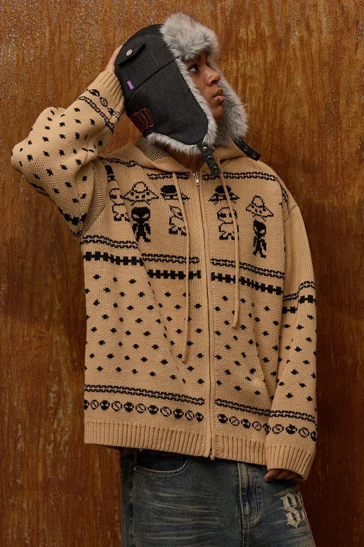 Person wearing beige zip-up knit hoodie with black alien and UFO pattern, and dark trapper hat with fur lining