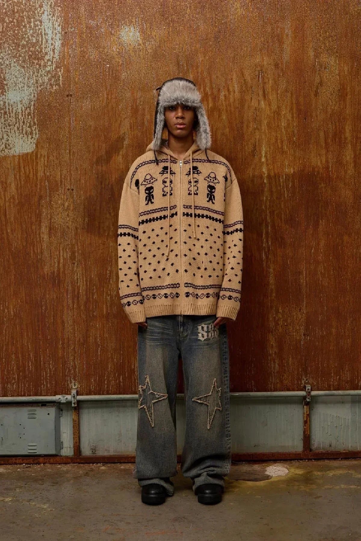 Model wearing beige alien pattern knit zip-up hoodie and loose denim jeans with star patches against rusted metal wall