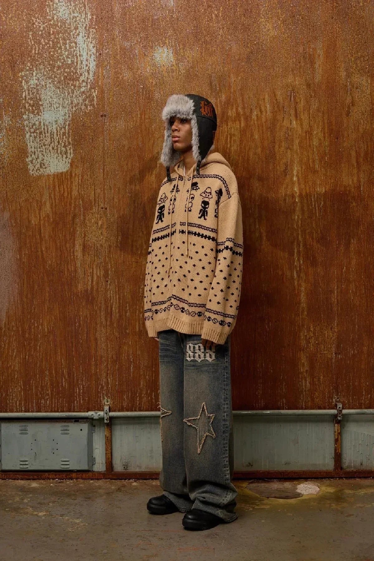 Person wearing beige alien pattern knit hoodie with drawstrings, gray star-patched jeans, black fur-trimmed trapper hat, and black shoes against rusty wall