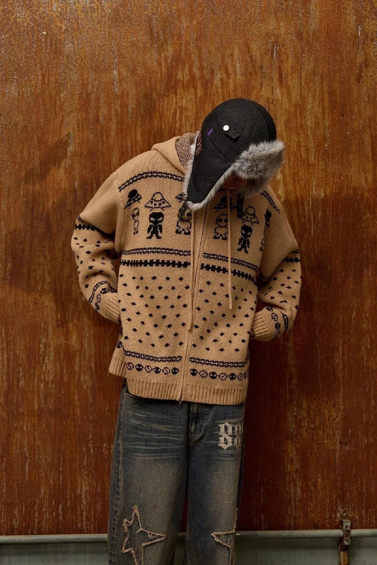 Beige knit zip-up hoodie with black alien pattern, worn with fur-lined black hat and distressed jeans