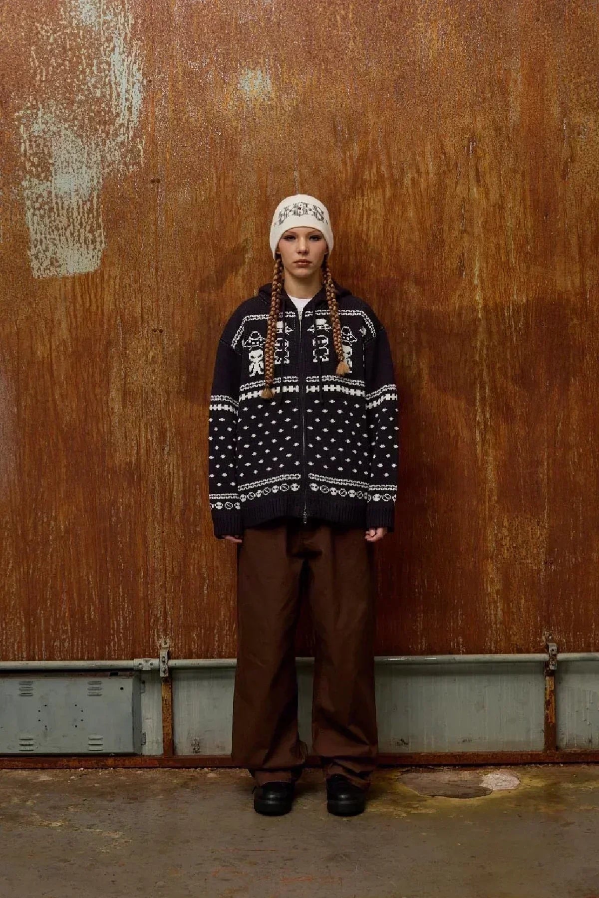 Model wearing navy zip-up knit hoodie with white alien and skull patterns, brown pants, and white skull beanie