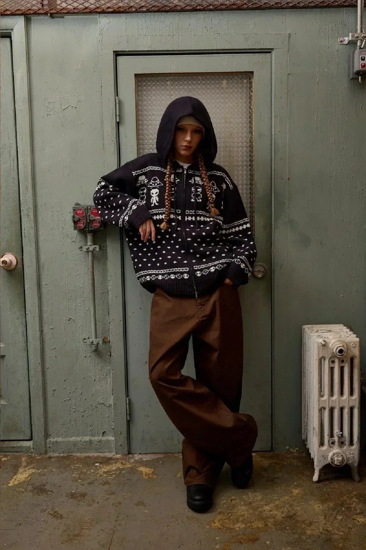 Model wearing navy alien pattern knit hoodie zip-up with brown wide-leg pants in an industrial setting