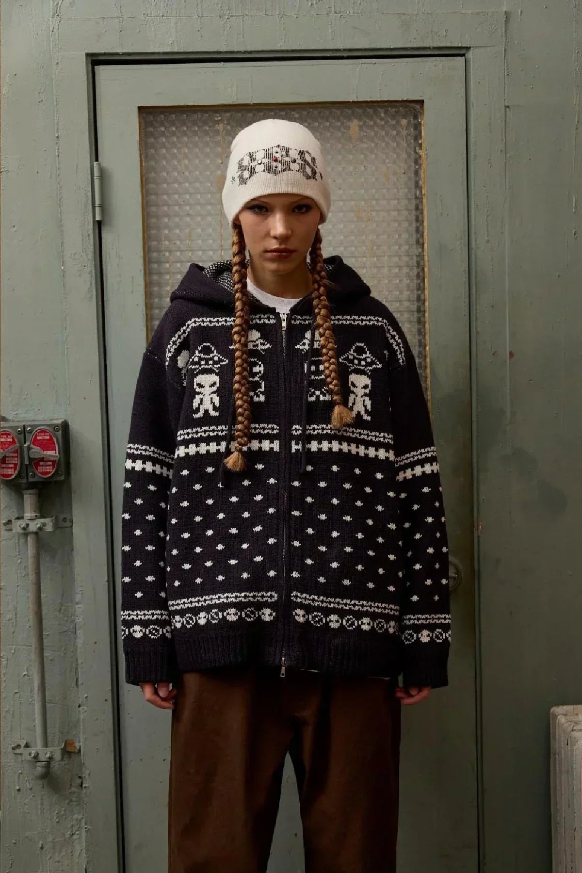 Model wearing navy blue zip-up knit hoodie with white alien and UFO pattern, paired with white beanie and brown pants