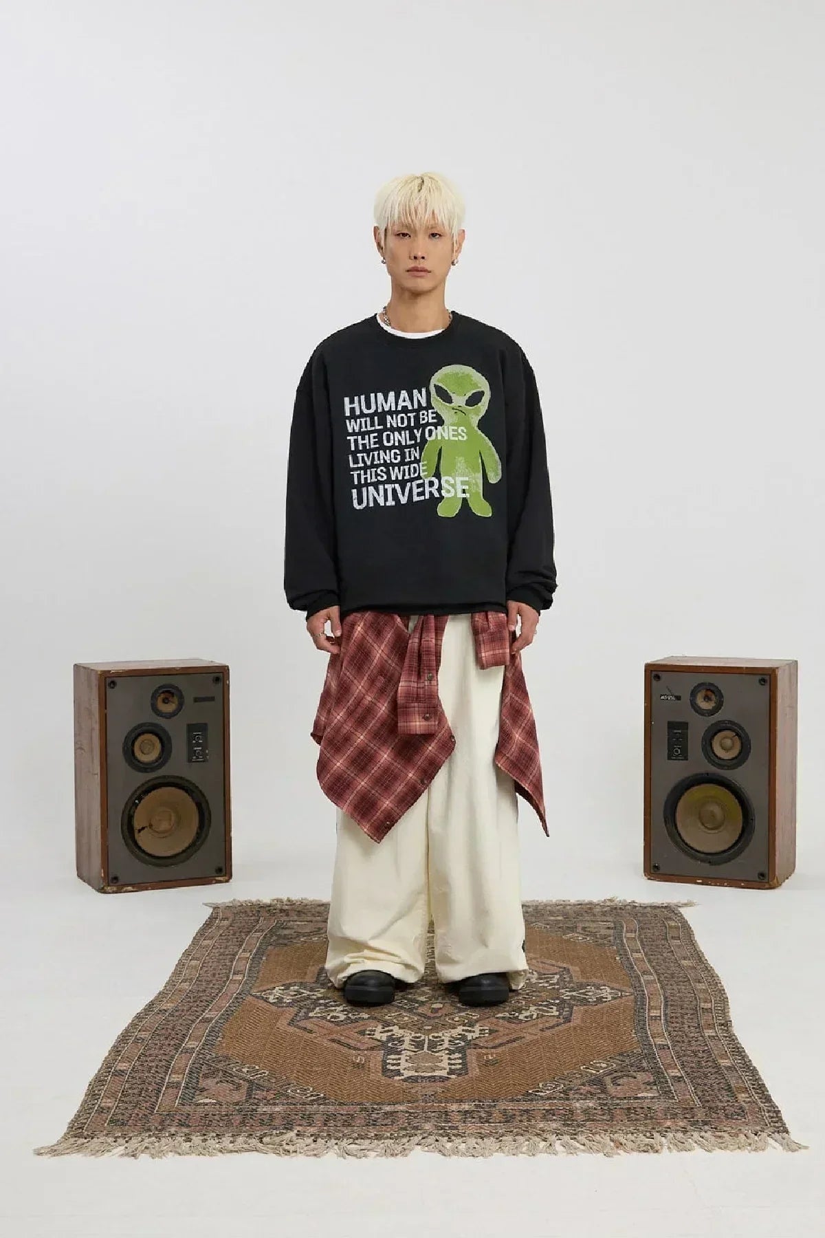 Young man wearing black alien slogan sweatshirt, beige wide pants, red plaid shirt tied around waist, standing on patterned rug