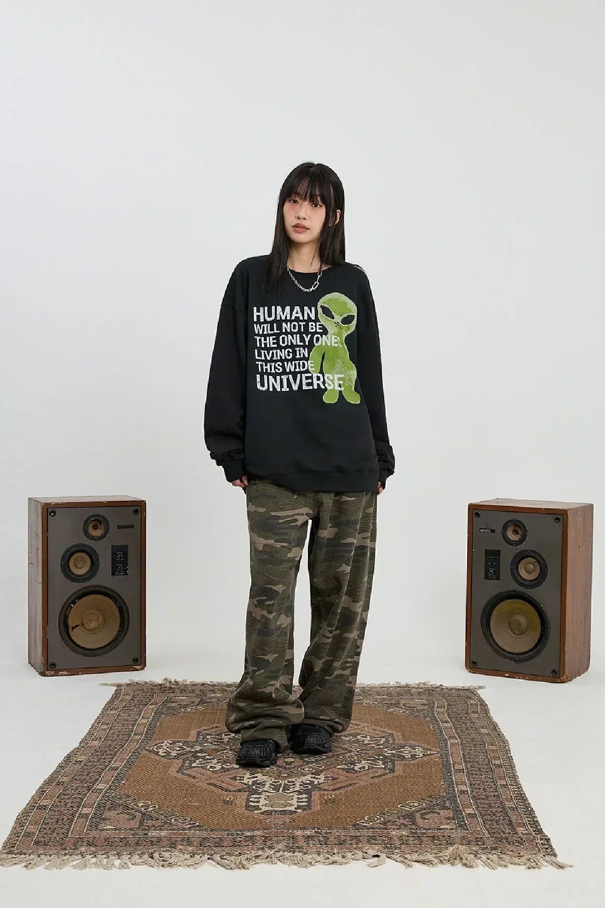 Woman wearing black alien slogan sweatshirt and camouflage pants standing on patterned rug between vintage speakers
