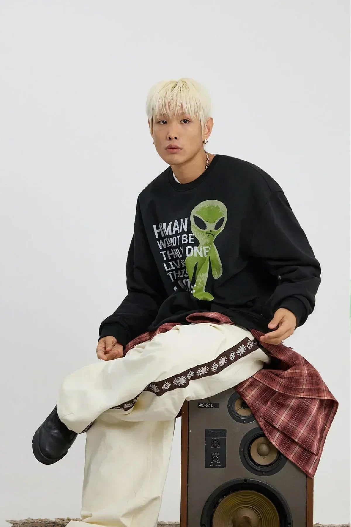 Young man with blonde hair wearing black alien graphic sweatshirt and white pants sitting on vintage speaker