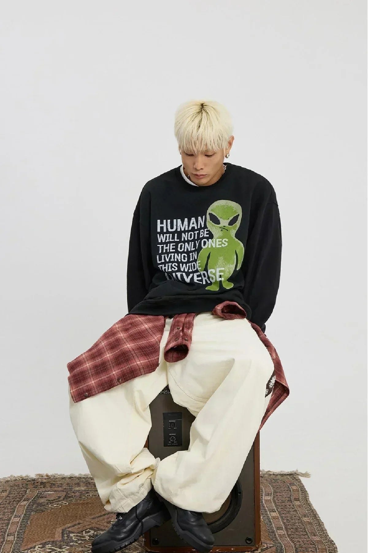 Young man with blonde hair wearing black alien slogan sweatshirt, cream baggy pants, and red plaid shirt sitting on speaker
