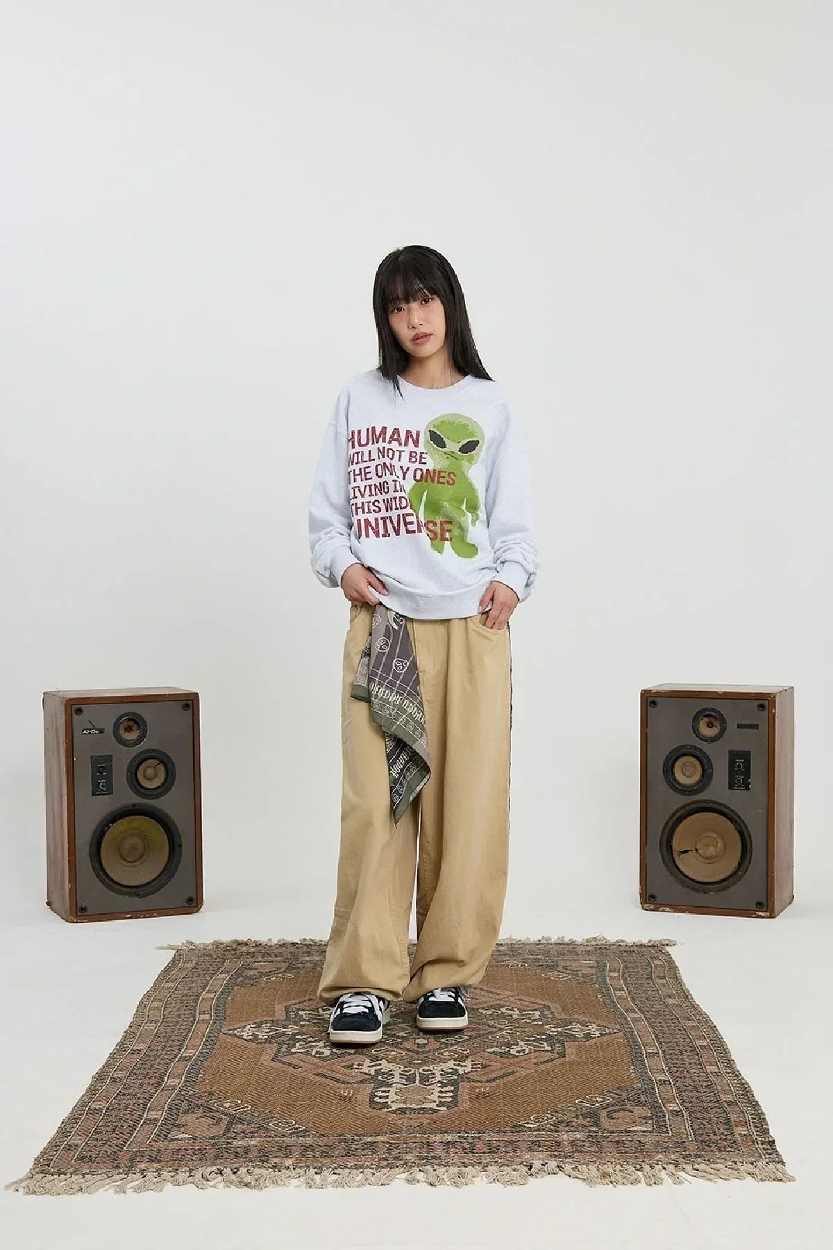 Woman wearing white alien slogan sweatshirt and beige wide pants standing on patterned rug between vintage speakers