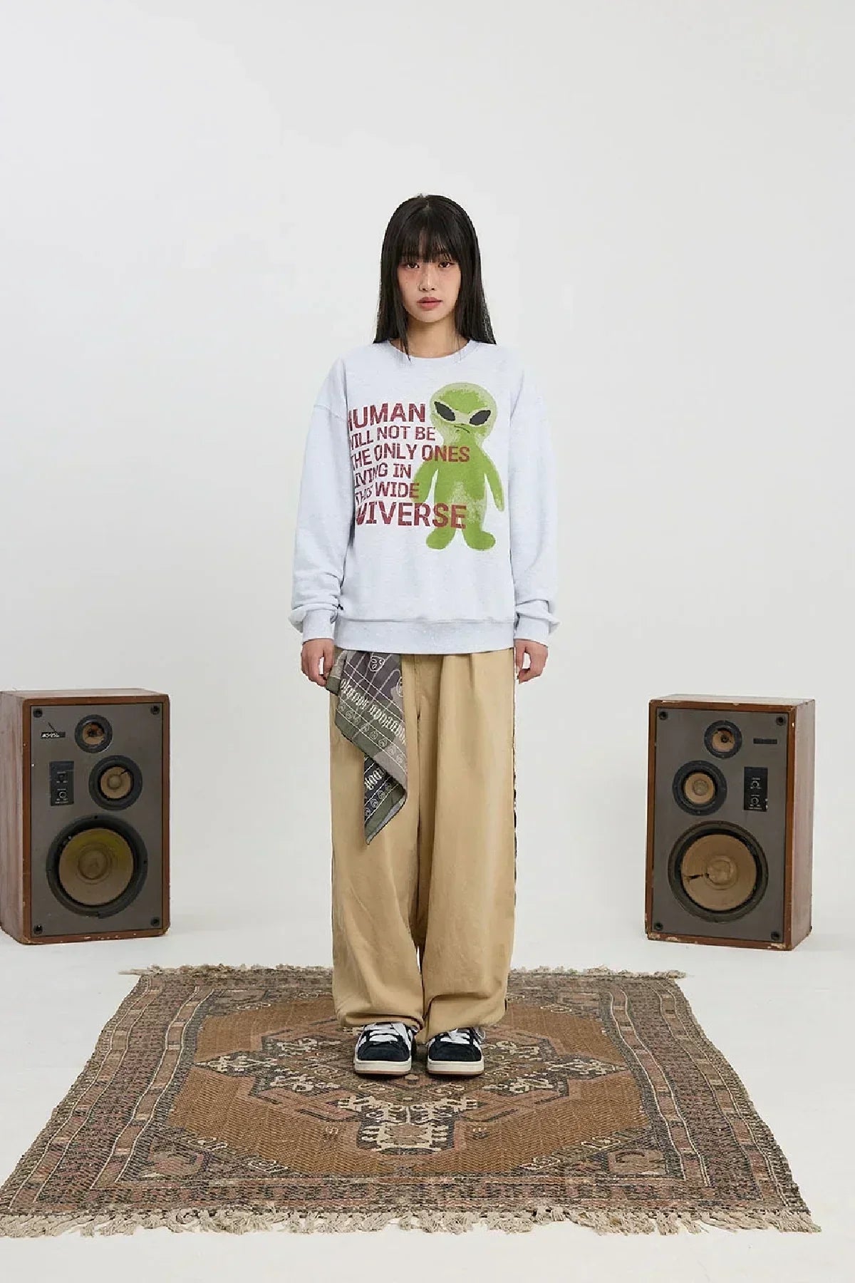 Young woman wearing melange white sweatshirt with green alien and slogan, wide khaki pants, standing on patterned rug between two speakers