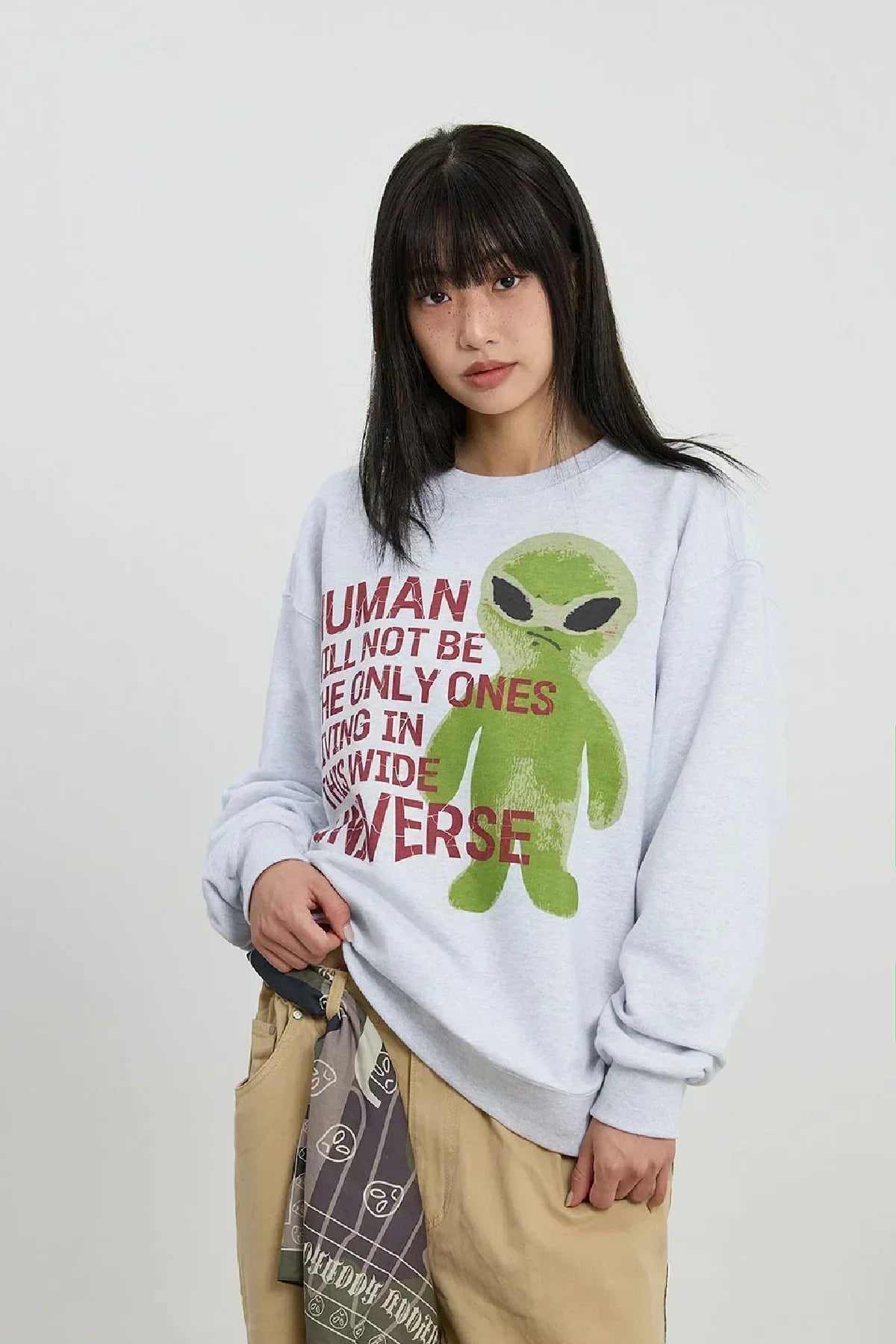 Model wearing light grey sweatshirt with green alien graphic and red text, beige pants, and bandana