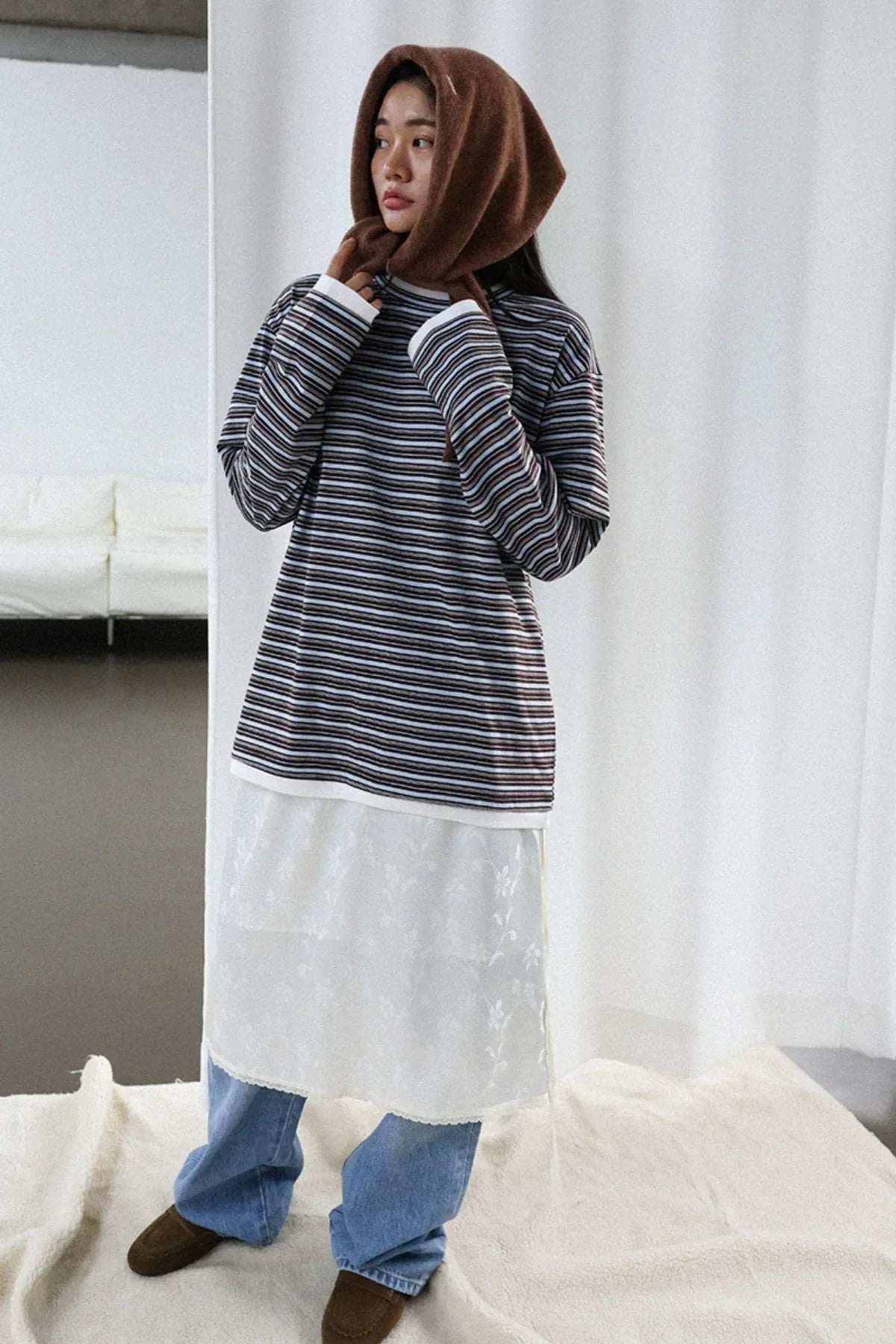 Woman wearing brown alpaca hooded muffler with striped top, white floral dress overlay, blue jeans, and brown shoes indoors