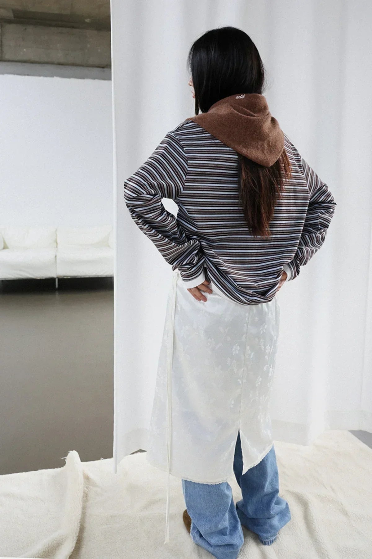 Woman wearing brown alpaca muffler, striped top, white skirt, and blue jeans in modern indoor setting