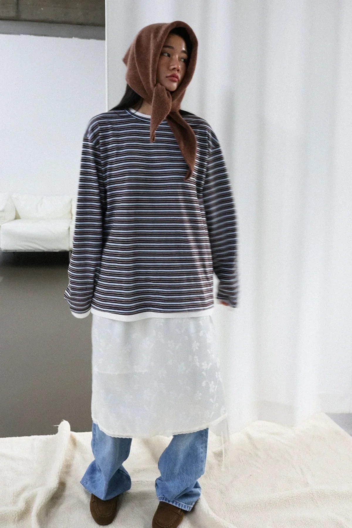 Model wearing brown alpaca scarf, striped long-sleeve shirt, white floral apron dress, blue jeans, and brown shoes in studio