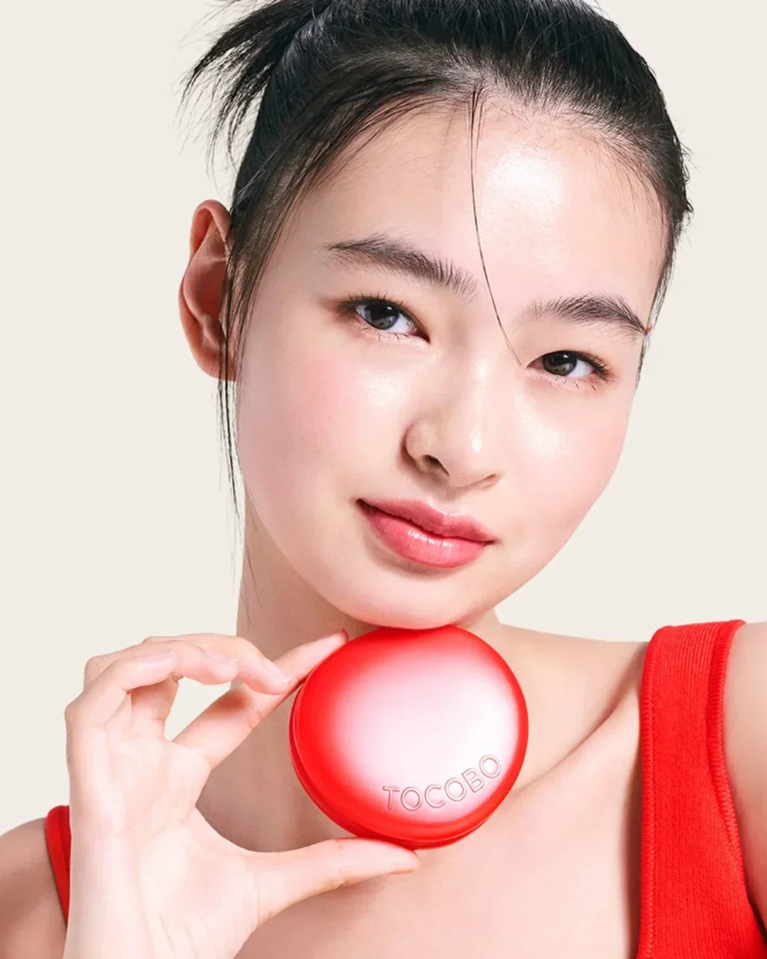 Young woman with glowing skin holding red TOCOBO cushion compact, wearing red top