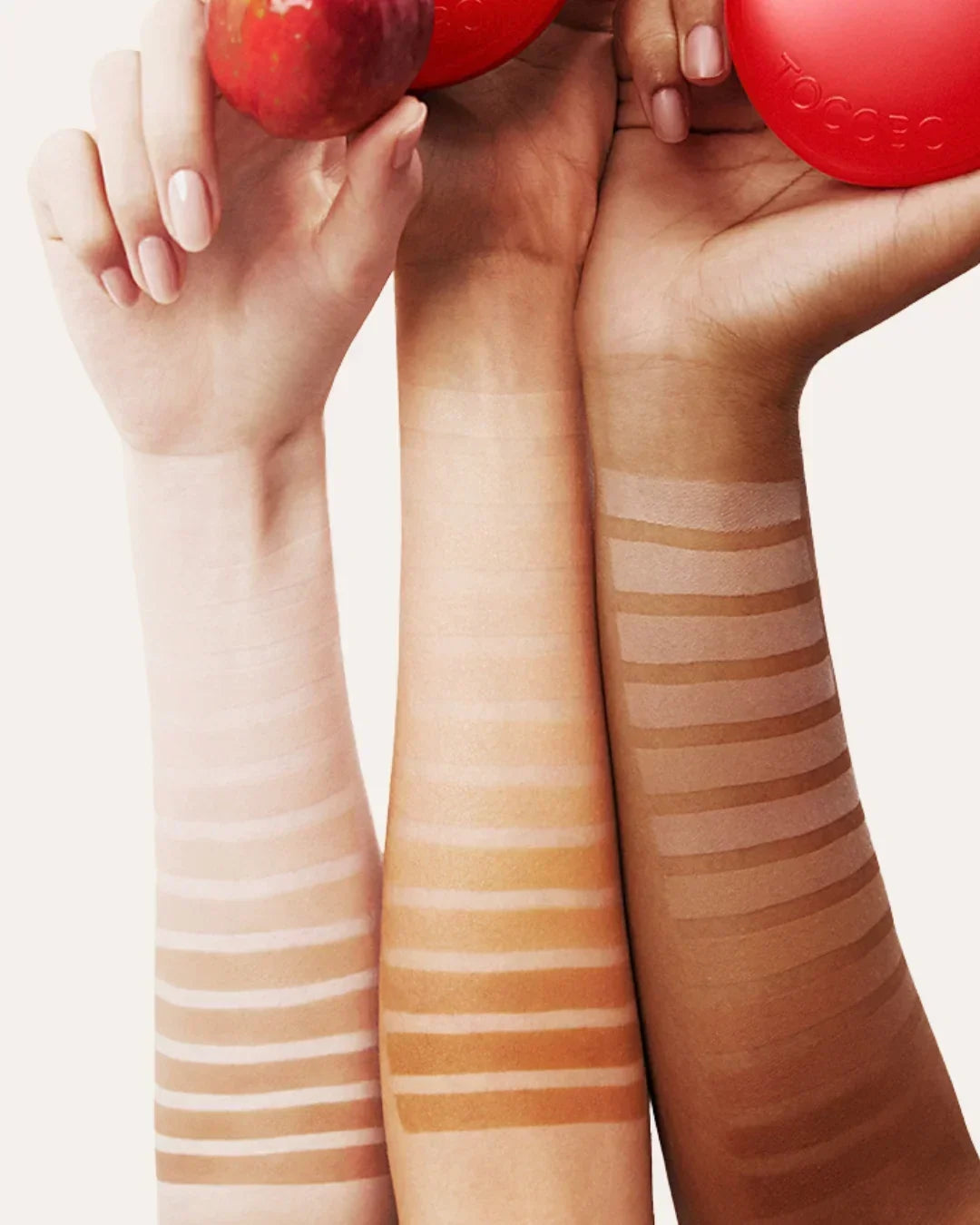 Three arms with diverse skin tones displaying swatches of makeup foundation shades in horizontal stripes