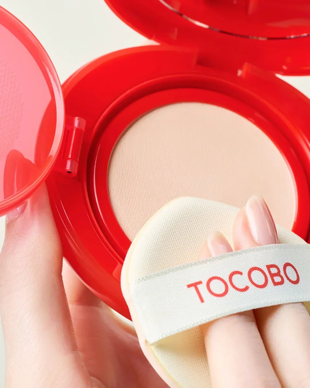 Hand holding red compact with beige TOCOBO cosmetic powder and applicator puff