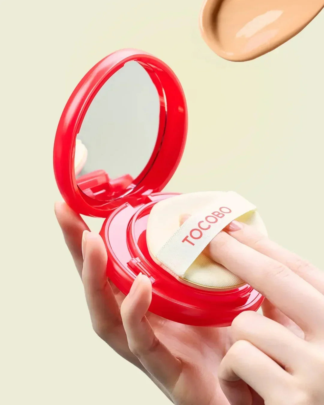 Hand holding red TOCOBO cushion compact with mirror and beige makeup sponge
