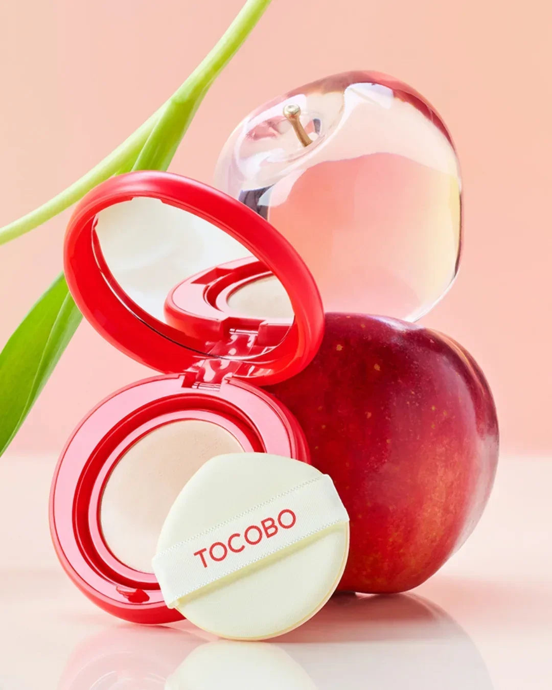 Tocobo Apple Dewy Fit Cushion compact with applicator next to a fresh red apple and glass apple-shaped decor