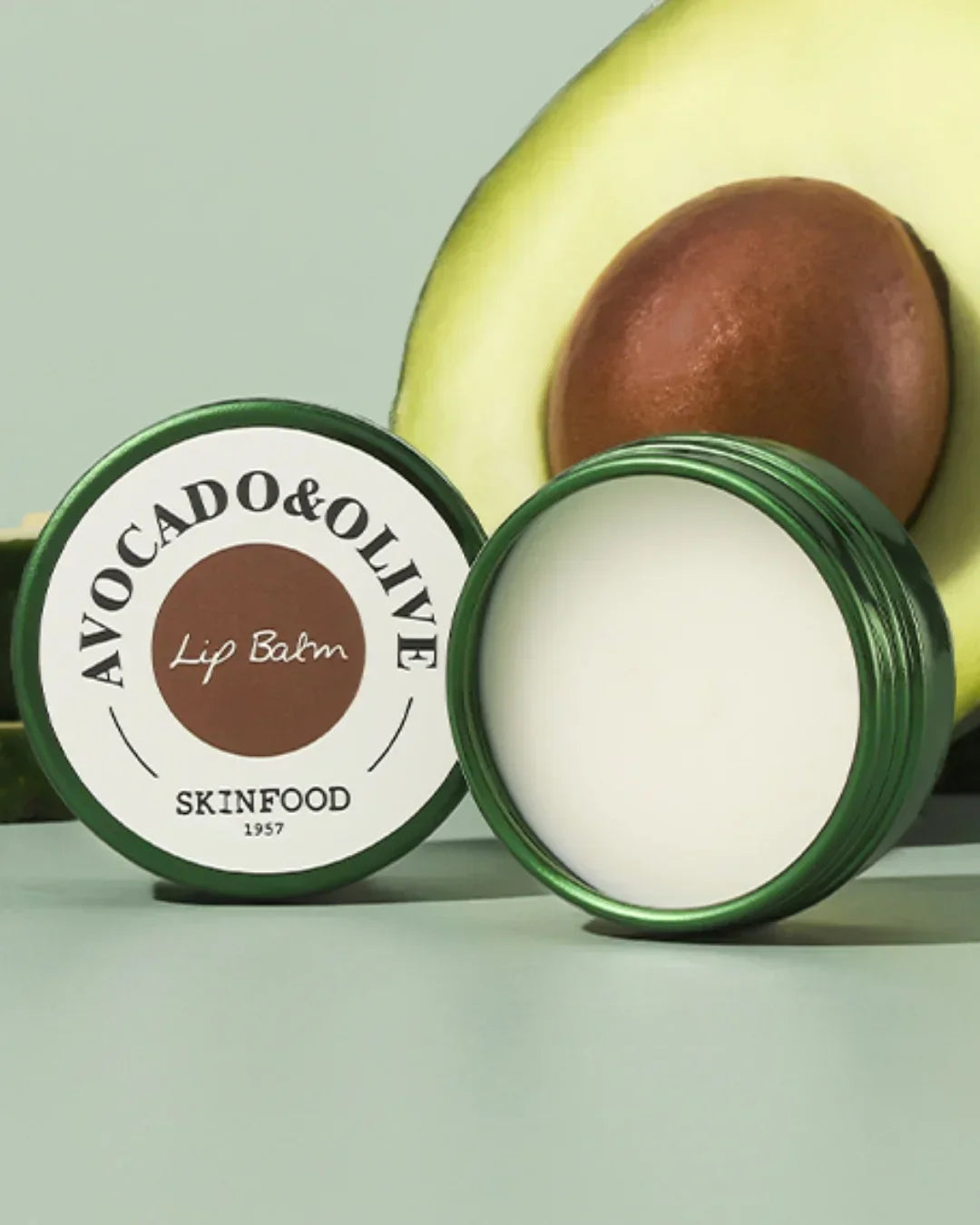 Open avocado & olive lip balm container with creamy texture and a halved avocado background