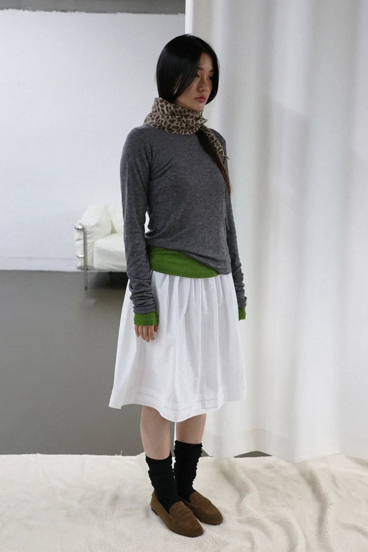 Woman wearing charcoal long sleeve shirt with green cuffs, white skirt, leopard print scarf, black socks, and brown loafers indoors
