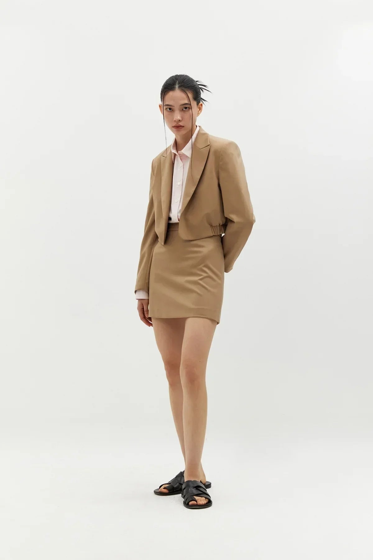 Young woman wearing beige cropped blazer and matching short skirt with black sandals in studio