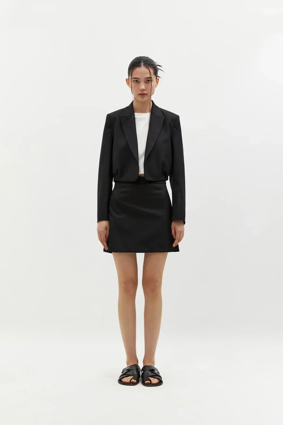 Woman wearing black wool mini skirt with black cropped blazer and white top, standing on white background