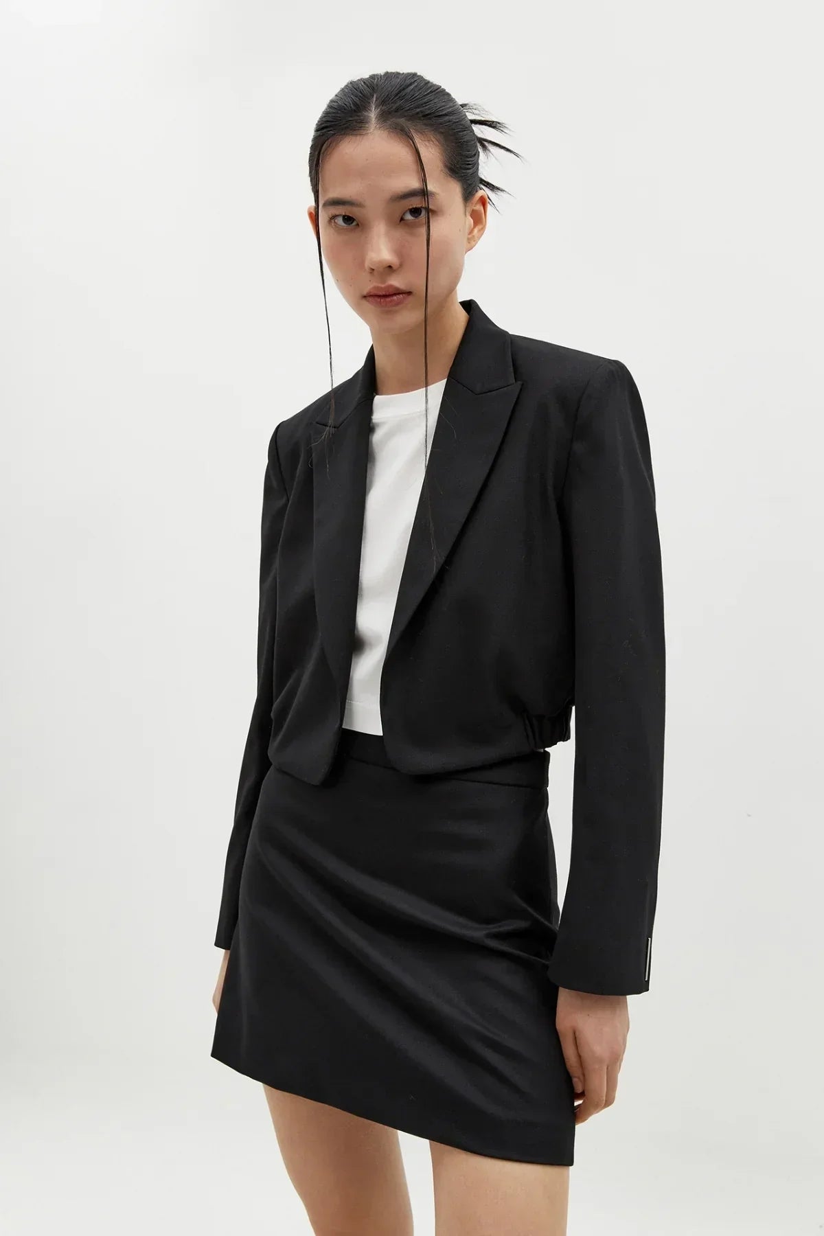 Model wearing a black cropped blazer and black mini skirt with a white top in a plain studio setting