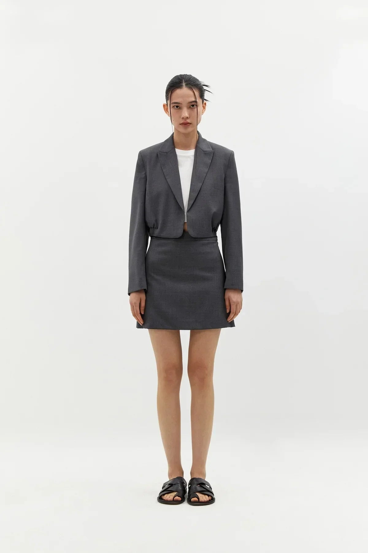 Woman wearing grey cropped blazer and matching wool mini skirt with white top and black sandals